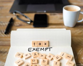 Closeup on notebook over vintage desk surface, front focus on wooden blocks with letters making Tax Exempt text. Business concept image with office tools and coffee cup in background